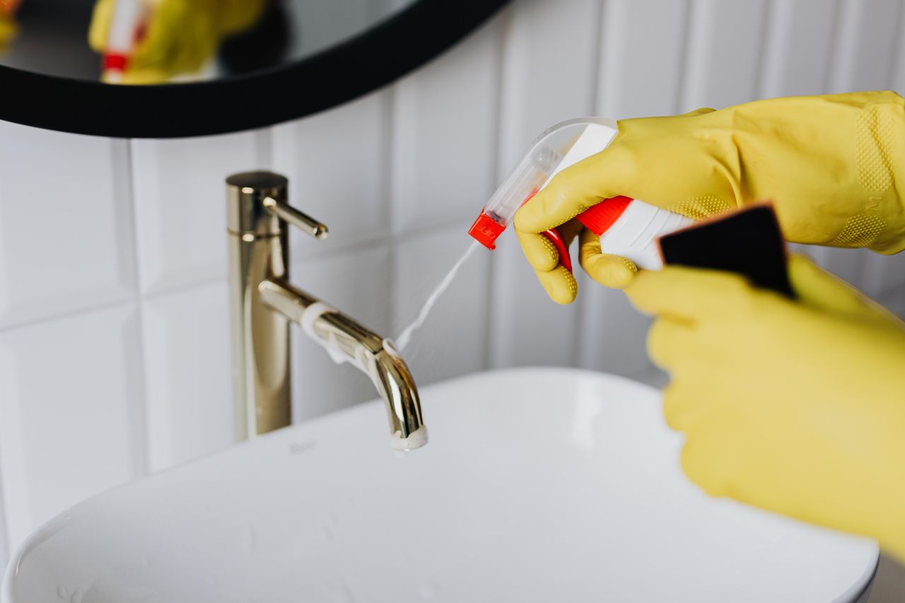 Seen from above, an anonymous housekeeper wearing yellow rubber gloves sprays cleaner onto a chrome bathroom water faucet